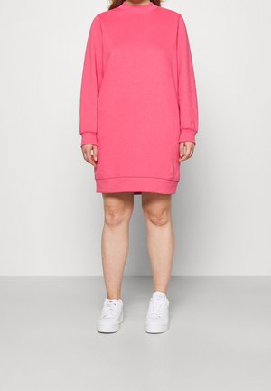 Personne porte une robe sweat rose à manches longues et longueur au genou, associée à des baskets blanches, debout devant un fond gris clair uni.