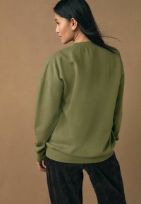 Sweat-shirt vert olive avec une coupe décontractée, poignets et ourlet côtelés. Détails incluant un motif subtil sur la manche. Porté avec un pantalon en velours côtelé foncé.