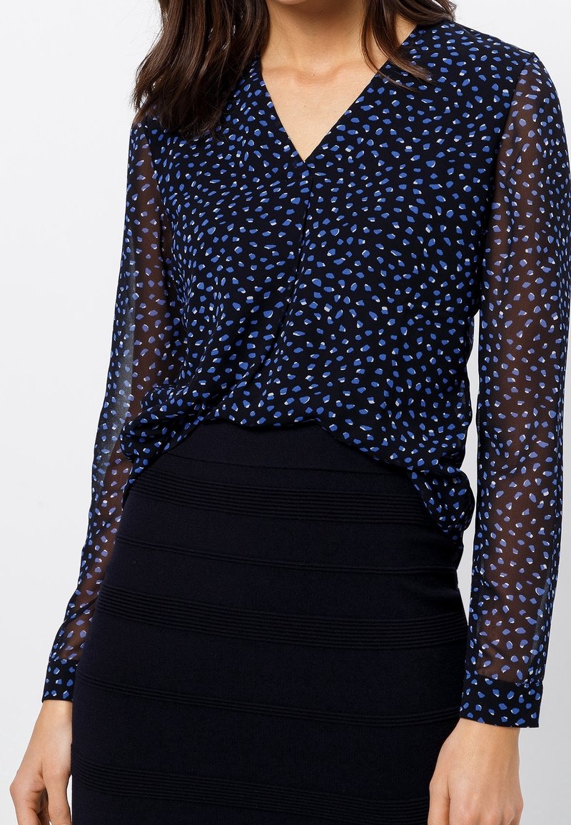Top nero con maniche lunghe trasparenti, caratterizzato da un motivo a pois blu e un design a scollo a V, abbinato a una gonna a tubino nera con texture.