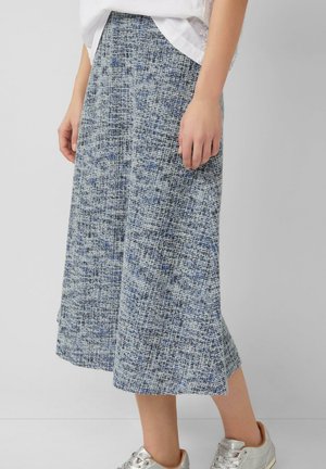 Frau trägt einen blau-weiß gemusterten Midirock, ein weißes Oberteil und silberne Sneakers, steht vor einem schlichten grauen Hintergrund.