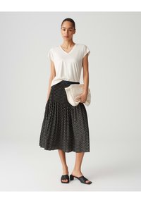 T-shirt blanc à col en V, jupe plissée noire avec motifs géométriques, sandales glissantes noires et sac à bandoulière crème.