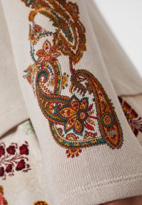 Tissu beige avec broderie détaillée de fleurs et motif paisley en fils orange, rouge, vert et jaune sur un textile tricoté.