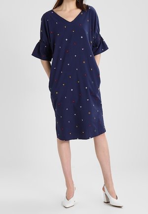 Žena v tmavě modrých šatech po kolena s barevnými malými vyšívanými tvary a bílými vysokými podpatky, stojící s rukama v kapsách.