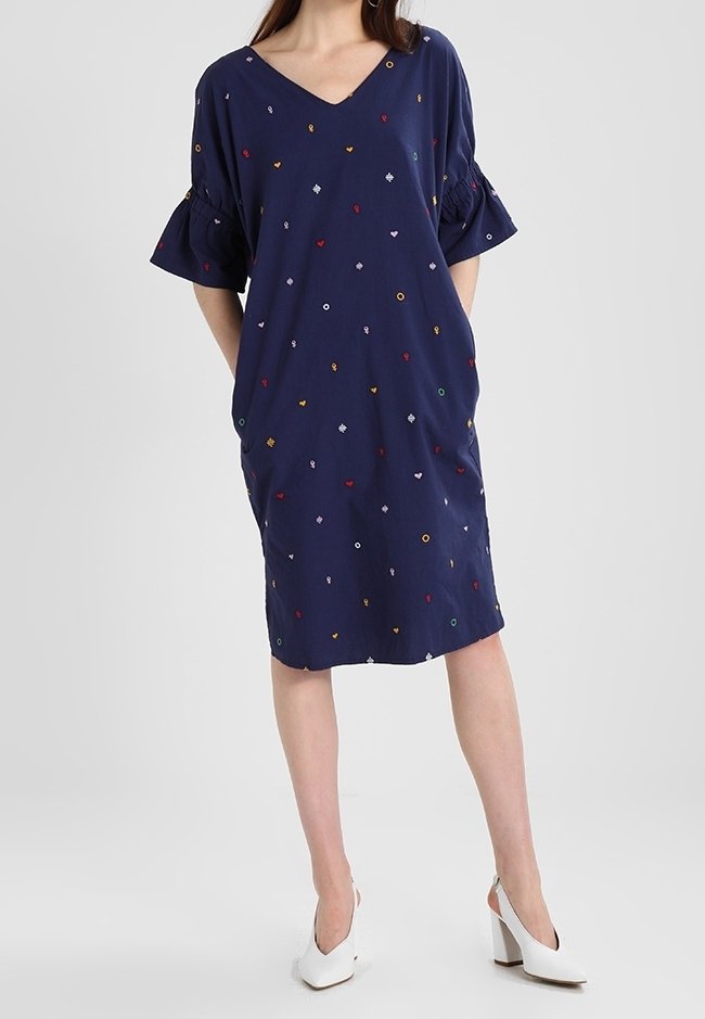 Femme portant une robe bleu marine mi-longue avec de petites formes brodées colorées et des chaussures blanches à talons hauts, debout les mains dans les poches.