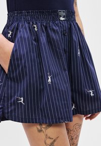 Shorts rayés bleu marine avec taille élastique, présentant des motifs de joueurs de tennis brodés en blanc. Comprend des poches avant et un détail de bouton.