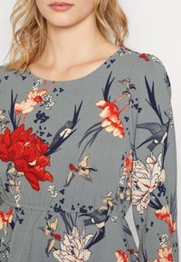 Kleid mit floralen Muster zeigt einen gedämpften grauen Hintergrund mit roten und cremefarbenen Blumen, blauen Akzenten und einem strukturierten Stoff. U-Bootausschnitt, taillierte Passform.