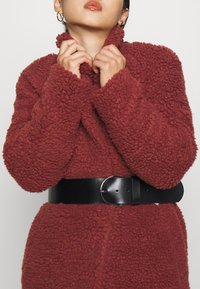 Röd, plyschig, texturerad kappa med hög krage, åtdragen vid midjan med ett svart läderbälte med en rund spänne. En mysig, tjock design.