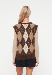 Brun og beige argyle-mønsteret vest med rund hals, strikket tekstur og ærmeløst design, over en beige langærmet skjorte, båret med en nederdel.
