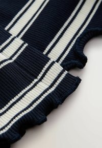 Tengerés ribbed pulóver részlet fehér és világosszürke csíkokkal, fodros szegéllyel. A közeli felvétel a textúrára és a színkontrasztra összpontosít.