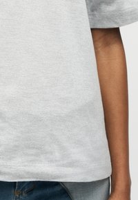 Άτομο που φοράει ανοιχτό γκρι κοντομάνικο πουκάμισο και μπλε τζιν παντελόνι, δείχνοντας τον κορμό και το δεξί χέρι σε απλό φόντο.