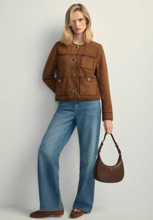 Blondynka w brązowej zamszowej kurtce zapinanej na guziki, niebieskich szerokich dżinsach, brązowych plecionych balerinach, trzymająca brązową skórzaną torebkę przy boku.