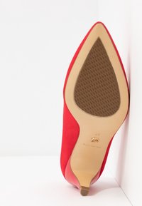 Chaussure à talon haut rouge à bout pointu montrant une semelle beige avec une zone d’adhérence texturée marron et une petite pointe de talon, en angle sur fond blanc.