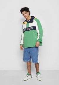 Zaļi un balti krāsu blokos veidots vējjaka ar ziliem akcentiem, ar logo joslu, apvienota ar ziliem džinsu šortiem un baltām kedām.