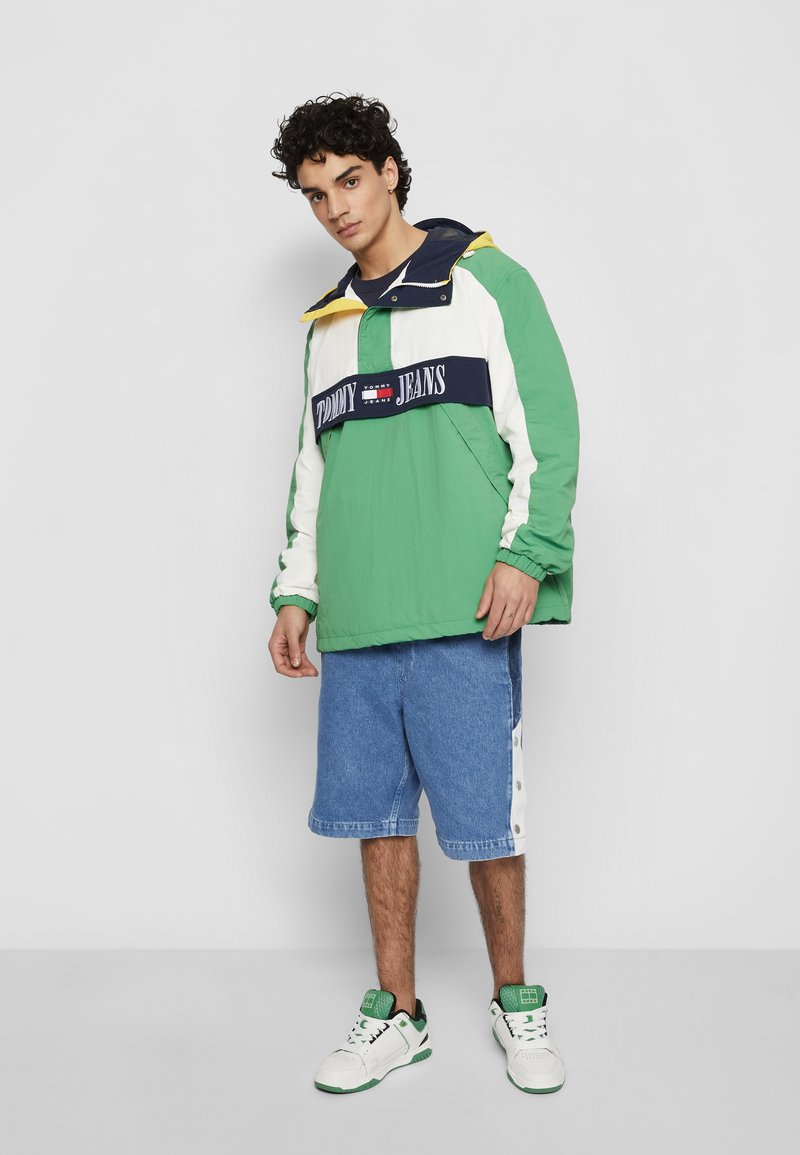 Zaļi un balti krāsu blokos veidots vējjaka ar ziliem akcentiem, ar logo joslu, apvienota ar ziliem džinsu šortiem un baltām kedām.