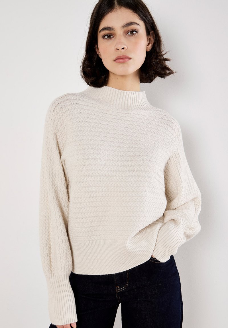 Sweterek w kolorze off-white, dziany, z wysokim prążkowanym kołnierzem, teksturowanym wzorem oraz prążkowanymi mankietami. W połączeniu z ciemnymi dżinsami, które mają luźny krój.