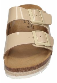 Beige patent leather slide sandal with two adjustable buckled straps, cushioned cork footbed, and textured rubber sole with jute detailing.