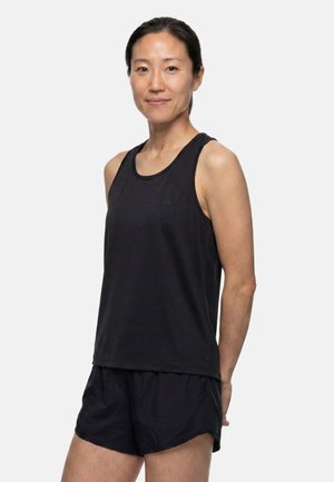 Femme portant un haut de sport noir sans manches et un short de sport noir, debout les mains dans le dos sur un fond uni.