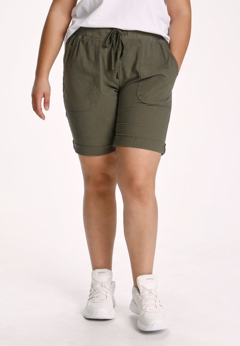 Person iført olivengrønne shorts med snøre og forlommer, hvid t-shirt og hvide Adidas-sneakers, stående foran en ensfarvet baggrund.