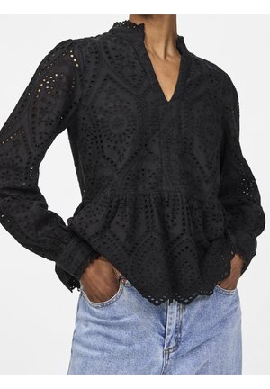 Blouse noire à manches longues en broderie anglaise avec bords festonnés et taille à volant, portée avec un jean bleu clair, mains dans les poches du jean.