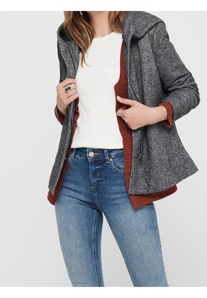 Kvinde iført lyseblå jeans, hvid top og åben grå hættejakke med rustfarvet foer, holder i jakke kanterne.