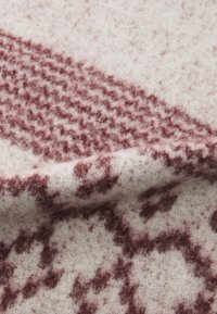 Couverture en laine douce avec un fond crème et des motifs abstraits bordeaux, surface texturée et finition légèrement duveteuse.