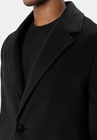 Zwarte jas van een wolmix met een ingesneden reverskraag, enkele knoopsluiting en een gestructureerde stof. Gedragen over een zwarte trui met een ronde hals.