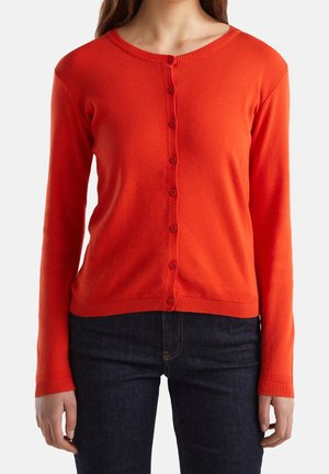 Femme portant un cardigan rouge vif boutonné à manches longues, associé à un jean bleu foncé, montrée du cou aux hanches.