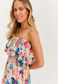 Blondynka w kolorowej, kwiatowej sukience z falbanami bez rękawów i naszyjniku z koralików, delikatnie uśmiechająca się, na jednolitym jasnym tle.