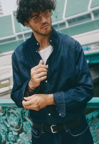 Camisa de denim azul marino de hombre con cuello abotonado, mangas largas y herrajes plateados. Se lleva con un cinturón negro, mostrando varios anillos en las manos.