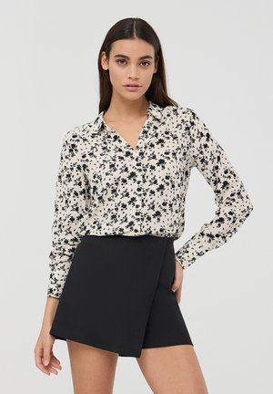 Kremowa bluzka w kwiaty z czarnymi wzorami, z kołnierzykiem i zapięciem na guziki, zestawiona z czarnymi szortami z wysokim stanem. Gładka faktura.