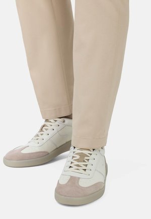 Witte leren sneakers met beige suède accenten en een lichtgrijze zool, gecombineerd met beige broek, en waarbij de veters en stikdetails zichtbaar zijn.