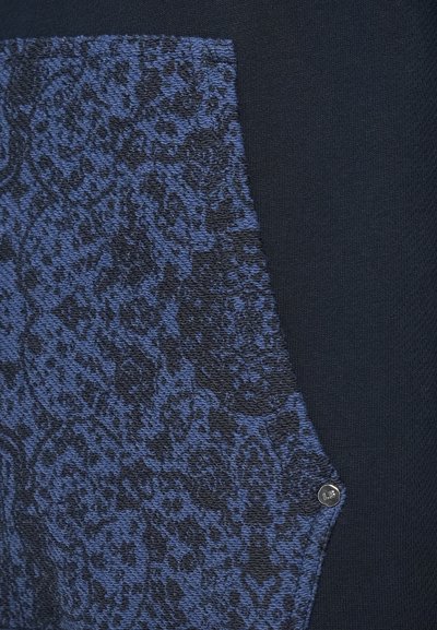 Marinepullover mit einem blauen strukturierten Einsatz und einem gemusterten Design, akzentuiert durch einen silbernen Logo-Button an der Seite.