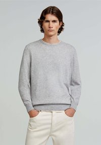 Pulover tricotat de culoare gri deschis, cu guler rotund, mâneci lungi și manșete cu cordon, purtat cu pantaloni de culoare crem. Textură netedă.