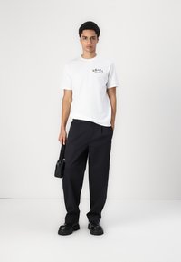 Hvid bomulds t-shirt med grafisk tryk, parret med løse sorte bukser og kraftige sorte sko, mens der holdes en lille sort taske.