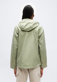 Άτομο που φοράει ανοιχτοπράσινο αδιάβροχο μπουφάν με κουκούλα και μπεζ παντελόνι cargo, εμφανιζόμενο από πίσω πάνω σε απλό λευκό φόντο.