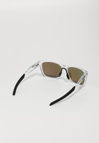 Klare rechteckige Sonnenbrille mit braunen getönten Gläsern, schwarzen Gummiohrstücken und transparentem Rahmen. Einfaches Design ohne zusätzliche Muster.