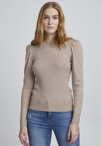 Beige ribbad långärmad topp med puffärmar, tillverkad av ett mjukt material. Bärs med blå slitna jeans och har en rund halsringning.
