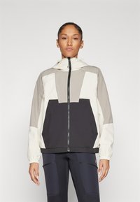 Chaqueta ligera con capucha que presenta una mezcla de colores beige, crema y negro, con cierre de cremallera frontal y un gran bolsillo en la parte delantera. Textura suave.