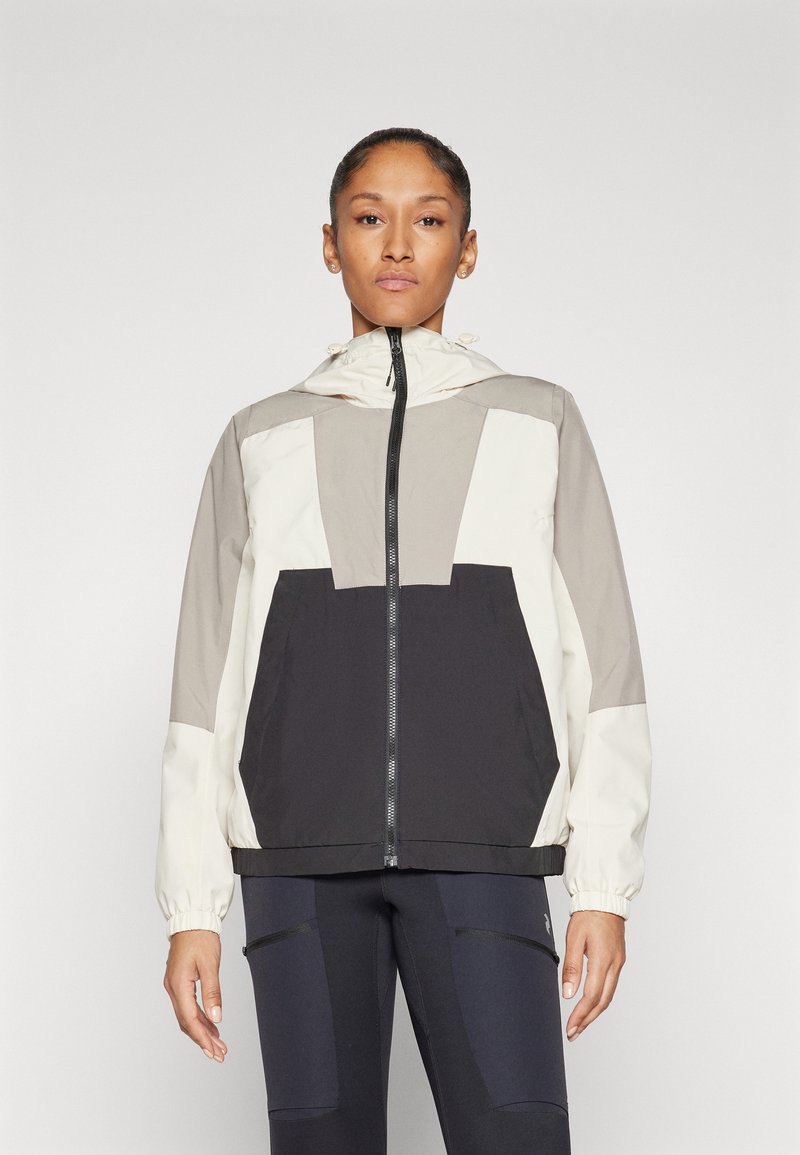 Chaqueta ligera con capucha que presenta una mezcla de colores beige, crema y negro, con cierre de cremallera frontal y un gran bolsillo en la parte delantera. Textura suave.