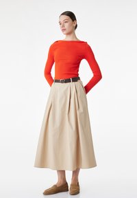 Mujer de pie con las manos detrás de la espalda, vistiendo una blusa de manga larga de color naranja brillante, falda midi beige con cinturón y mocasines marrones sobre fondo blanco.