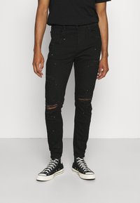 Jeans de mezclilla negros con detalles rasgados, corte ajustado y salpicaduras de pintura dispersas. Combinados con zapatillas altas negras.