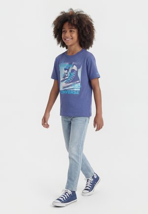 Enfant souriant aux cheveux bouclés, portant un t-shirt graphique Converse bleu, un jean bleu clair et des baskets montantes bleu marine.