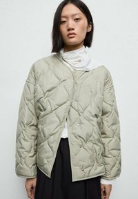 Giacca leggera imbottita verde chiaro con bottoni, caratterizzata da una vestibilità ampia e un motivo a rombi, indossata sopra una camicia bianca con colletto.