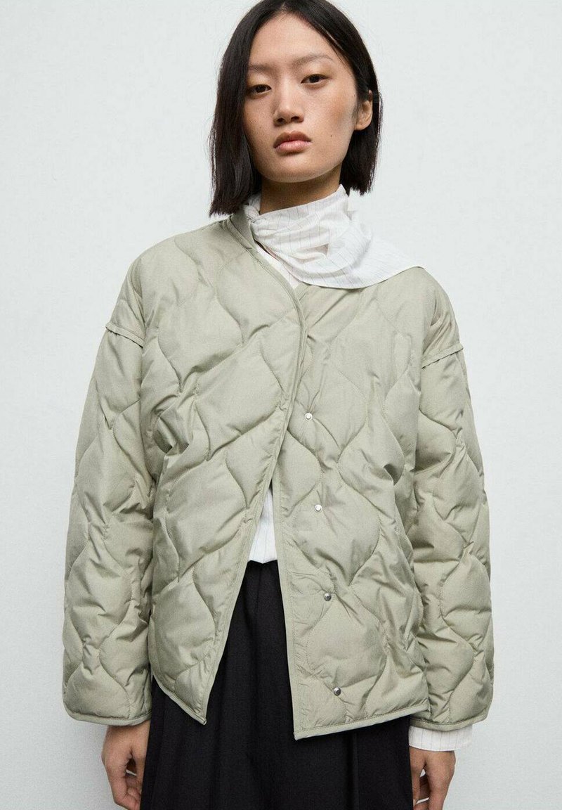 Giacca leggera imbottita verde chiaro con bottoni, caratterizzata da una vestibilità ampia e un motivo a rombi, indossata sopra una camicia bianca con colletto.