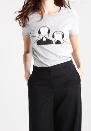 Žena nosící šedé tričko s černým motivem osoby a kočky, kteří oba mají sluchátka spojená jedním kabelem.