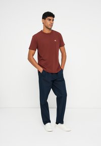 Μαροκό βαμβακερό μπλουζάκι με στρογγυλή λαιμόκοψη και μικρό λογότυπο, σε συνδυασμό με ναυτικές παντελόνες και λευκά αθλητικά παπούτσια. Το μοντέλο στέκεται με τα χέρια στις τσέπες.