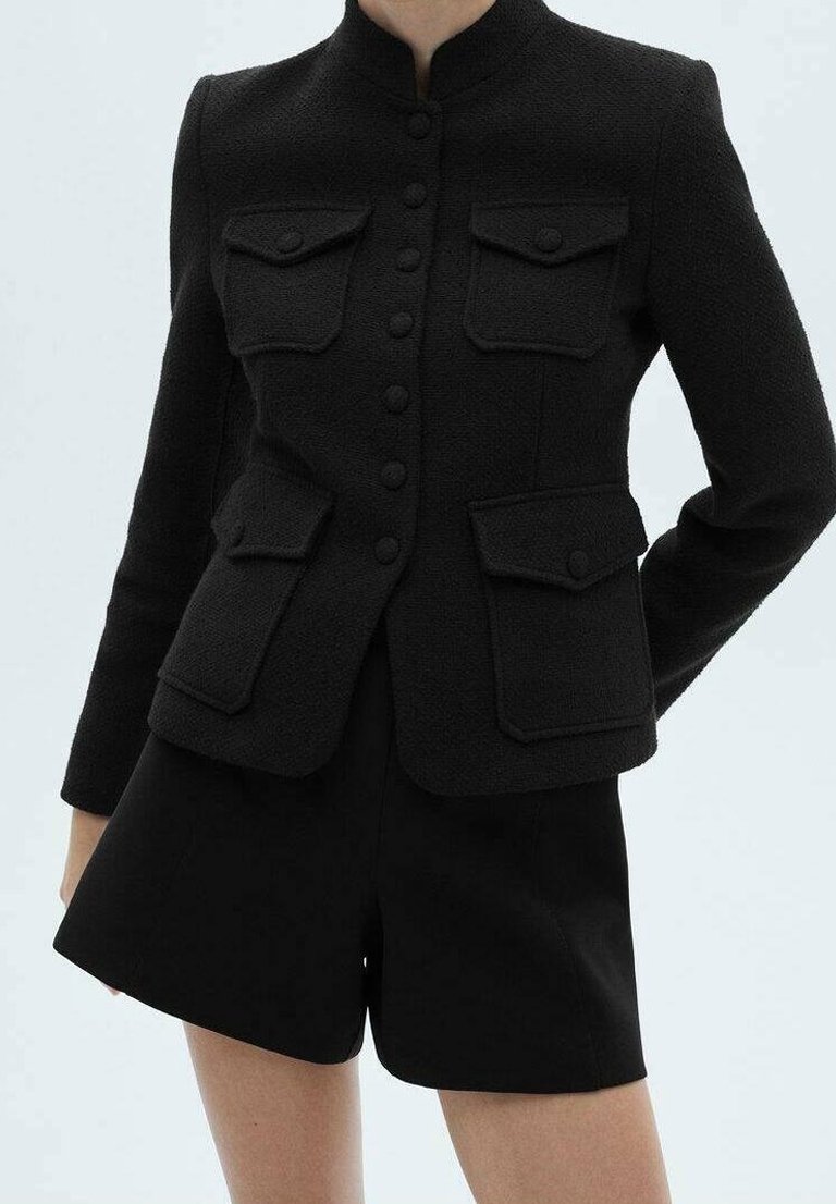 Femme portant une veste noire texturée ajustée avec quatre poches à rabat et un short noir assorti sur un fond uni.
