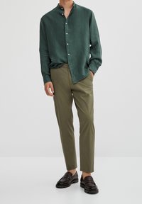 Mężczyzna w ciemnozielonej koszuli z guzikami, oliwkowych spodniach i ciemno brązowych mokasynach, stoi z jedną ręką w kieszeni.