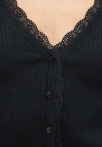 Μαύρη ριμπ ζακέτα με V-λαιμόκοψη διακοσμημένη με δαντέλα. Έξι μαύρα κουμπιά στο μπροστινό μέρος και λεπτές υφασματικές λεπτομέρειες σε όλη την επιφάνεια.