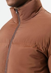 Bruine geïsoleerde jas met een hoge kraag, lichtgewicht stof en een ritssluiting. Heeft een gladde textuur en een aansluitend ontwerp.